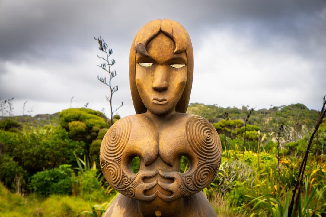 A carved wooden Māori pou with pāua-shell eyes standing among coastal bush